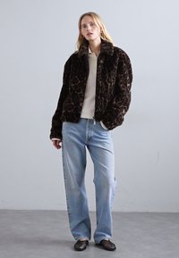 Woman wearing a dark leopard print faux fur jacket, white shirt, light blue jeans, and black woven slip-on shoes standing indoors.