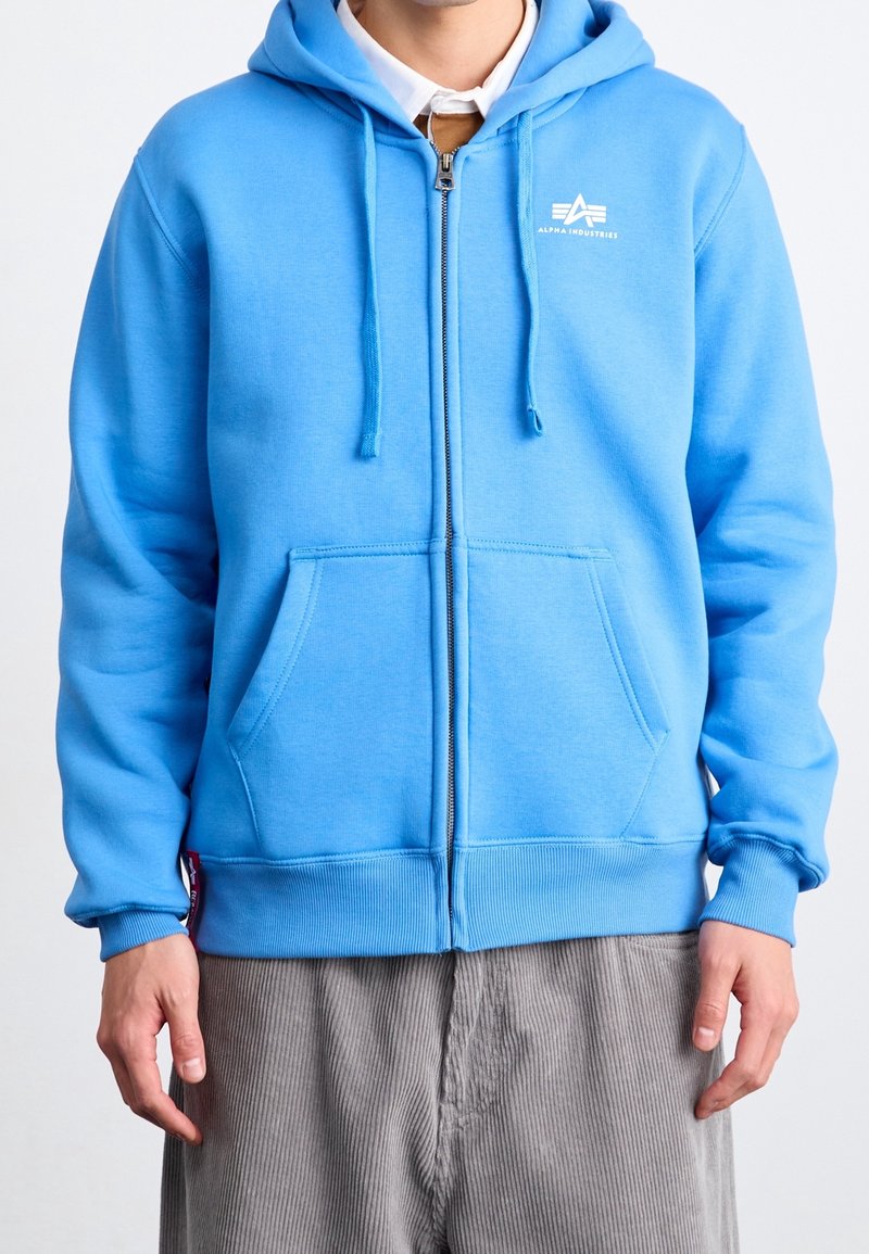 Homme portant un sweat à capuche zippé bleu clair avec des poches devant et le logo blanc Alpha Industries sur la poitrine, pantalon en velours côtelé gris visible.