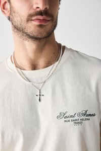 T-shirt beige clair avec logo en texte vert. Deux colliers superposés : un collier argenté avec un pendentif croix et un collier en perles argentées.