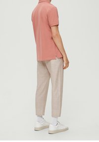 Man wearing a dusty pink short-sleeve polo, beige cropped pants, white socks, and white sneakers, standing against a plain background.