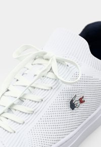 Hvite strikkede joggesko med teksturert mesh-design, utstyrt med flate hvite lisser og en liten brodert logo på siden.