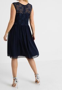 Robe bleu marine avec un corsage en dentelle et une jupe en chiffon fluide. Portée avec des chaussures à talons hauts argentées, montrant une texture lisse.