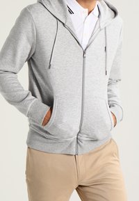 Grå zip-up hoodie med dragsko, som har kengurufickor, strukturerad tyg och en avslappnad passform, ihop med en vit polo tröja.