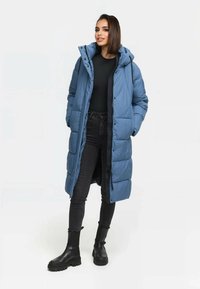 Długi, niebieski płaszcz puchowy z kapturem, z zamkiem błyskawicznym i zapięciem na napy, dwiema przednimi kieszeniami i pikowaną fakturą. Połączony z czarnymi dżinsami i butami.