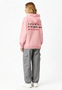 Felpa con cappuccio rosa con testo nero e rosso sul retro, abbinata a pantaloni cargo grigi e sneakers bianche. Look casual e vestibilità rilassata.