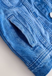 Kék farmerdzseki ujja szabályos hasíték zsebével és látható varrási részletekkel világos háttéren.