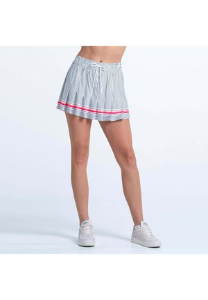 Frau trägt einen plissierten, weißen Minirock mit schwarzen vertikalen Streifen und roten horizontalen Akzenten, kombiniert mit weißen Turnschuhen, steht mit gespreizten Beinen.