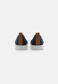 Námornické plátěné tenisky s hnědými koženými patkami, bílými gumovými podrážkami a minimalistickým designem. Zaoblený tvar a hladká textura.
