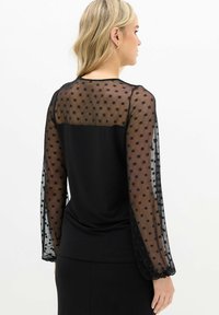 Vrouw met blond haar die een zwarte top draagt met doorschijnende lange mouwen met stippen en een effen zwarte achterkant, zijwaarts gekeerd tegen een witte achtergrond.