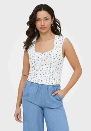 Woman with dark wavy hair wearing a white sleeveless top with blue floral print and light blue high-waisted pants, hand in pocket.
