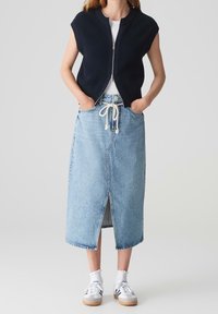 Kvinna som bär en ärmlös svart zip-front väst, vit skjorta, ljusblå denim midi kjol med framfåra och repbälte, vita strumpor och sneakers.