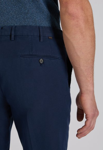 Zbliżenie na dolną część pleców i prawy ramię osoby noszącej granatowe spodnie z zapinanymi na guziki tylnymi kieszeniami oraz niebieską, teksturowaną koszulę.