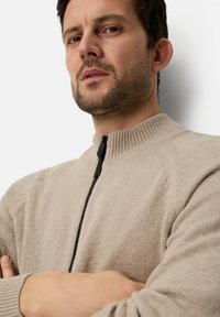 Beige zip-up sweater with textured knit, ribbed collar, and black zipper detail. Features raglan sleeves and relaxed fit.