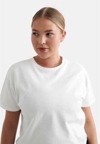 Vit bomulls t-shirt med korta ärmar och rund hals. Har en subtil präglad logotyp vid kragen. Slät textur över hela plagget.