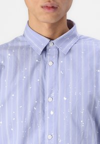 Light blue striped shirt with white paint splatters, button-down collar, and five buttons visible on the front. Cotton fabric, casual design.