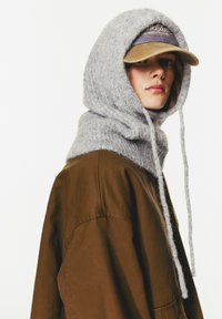 Grau gestrickte Kapuze mit langen Bändern, kombiniert mit einer braunen, oversized Jacke und einer khakifarbenen Mütze mit gesticktem Text. Weiche, strukturierte Materialien.