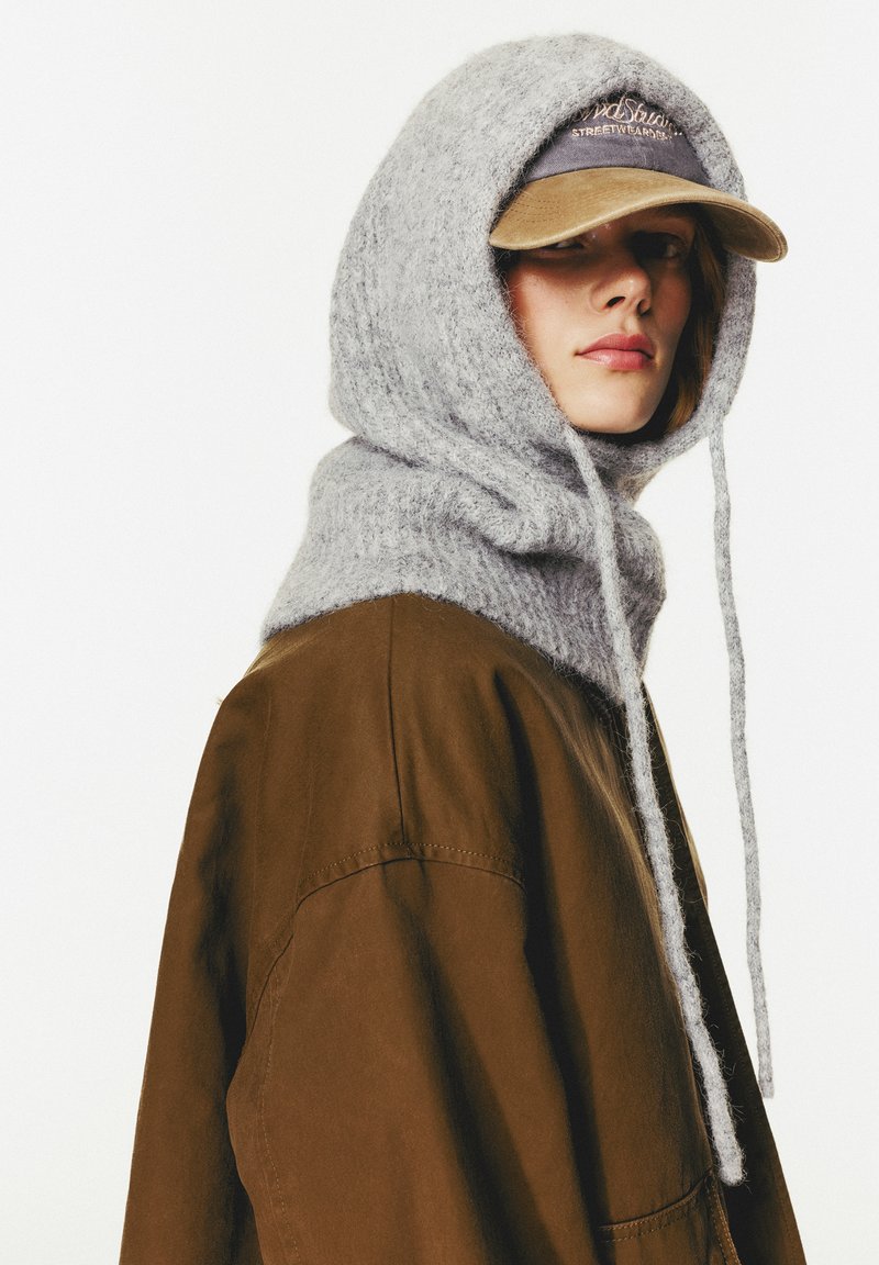 Grau gestrickte Kapuze mit langen Bändern, kombiniert mit einer braunen, oversized Jacke und einer khakifarbenen Mütze mit gesticktem Text. Weiche, strukturierte Materialien.
