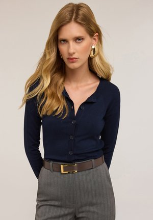 Cardigan navy con bottoni, caratterizzato da una texture a coste e colletto, indossato sopra dei pantaloni a righe grigie, fissato con una cintura marrone con fibbia dorata.