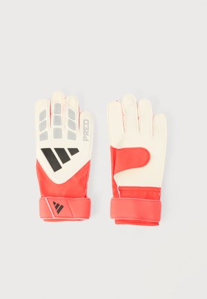 Punaste ja valgete värvidega väravavahi kindad musta Adidas logoga, pehmendatud sõrmedega ja reguleeritavate randmerihmadega, asetatud tasasele heledale taustale.