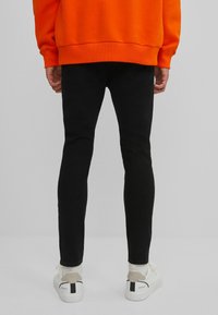 Černé skinny džíny, bavlněný materiál, hladká textura, v kombinaci s bílými teniskami s černými akcenty, nošeny s oranžovou mikinou.