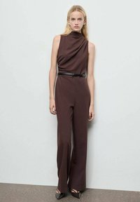 Bruine, mouwloze jumpsuit met een gedrapeerde halslijn, een ceintuur in de taille en een wijd broekontwerp. Glad materiaal met een subtiele glans en een getailleerde pasvorm.