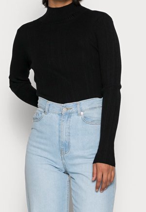 Persoon draagt een geribde zwarte coltrui met lange mouwen, ingestopt in een lichtblauwe jeans met hoge taille tegen een effen achtergrond.