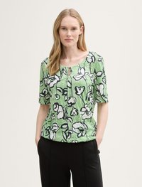 Grünes Oberteil mit floralem Druck und kurzen Ärmeln, mit rundem Halsausschnitt, schwarzem Blumenumriss und strukturiertem Stoff. Kombiniert mit schwarzen Hosen.