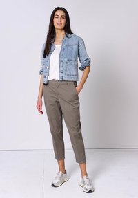 Femme debout portant une veste en jean bleu clair, un t-shirt blanc, un pantalon court kaki et des baskets blanches sur fond uni.