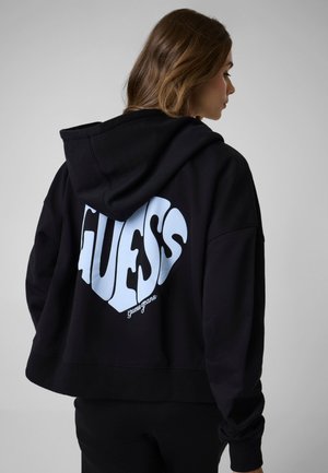 Vrouw met bruin haar die een zwarte hoodie draagt met een groot wit gestileerd "GUESS" logo op de rug in hartvorm.