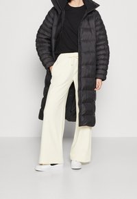 Svart puffjacka med lång silhuett och dragkedja fram. Bärs över en svart t-shirt och krämfärgade vida byxor, kompletterade med vita sneakers.