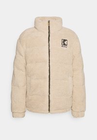 Giacca in pile beige con finitura testurizzata, dotata di un colletto alto, chiusura con zip e un piccolo logo nero sul lato sinistro del petto.