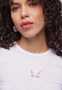 T-shirt bianco a coste con un design di farfalla ricamato in bianco sul petto. Modella con capelli ricci indossa orecchini a cerchio argentati.