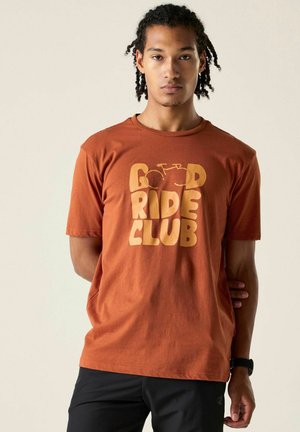 Młody mężczyzna z plecionymi włosami, ubrany w rdzawe t-shirt z napisem "Good Ride Club" i grafiką roweru, stoi z rękami za plecami.