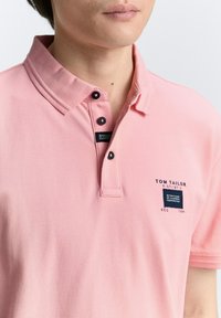 Rosa pikétröja med en tvåknapprad, broderad logotyp och taggdetaljer. Tyget har en strukturerad yta.