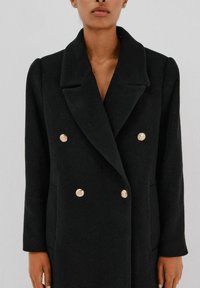 Mustat kaksirivinappinen takki, joka on valmistettu teksturoidusta kankaasta. Takissa on leveä kaulus ja edessä neljä metallista nappia.