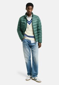 Grøn quilted jakke over en cremefarvet cable-knit sweater med blå accenter, parret med lyseblå jeans og hvide sneakers med sorte detaljer.