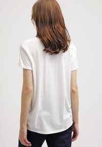 Bílé tričko s krátkým rukávem, zaobleným lemem a volným střihem. Měkký bavlněný materiál, hladká textura a detail s ohrnutými rukávy.