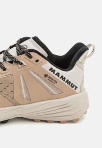 Chaussure de randonnée beige avec des accents noirs et blancs. Elle présente des panneaux en maille texturée, un col rembourré et un logo bien visible. Équipée de GORE-TEX.