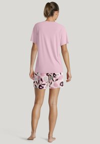Rosa T-Shirt mit kurzen Ärmeln kombiniert mit Shorts im Blumenmuster, die rosa, grüne und dunkelviolette Farbtöne vor einem hellrosa Hintergrund zeigen.