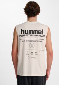 Ärmlös krämfärgad tanktop som har en stor svart logotyp på baksidan, vårdsymbolen och texten för "hummel PERFORMANCE" och "AUTHENTIC SPORTSWEAR."