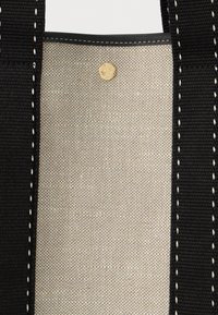 Μπεζ υφαντός καμβάς με χρυσό κουμπί σnap ανάμεσα σε δύο μαύρες λωρίδες με λευκή ραφή σε μια τσάντα.