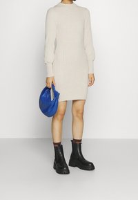 Beżowa sweterkowa sukienka z prążkowanym wzorem i długimi rękawami, zestawiona z czarnymi masywnymi botkami za kostkę i niebieską torebką. Prosty i dopasowany krój.