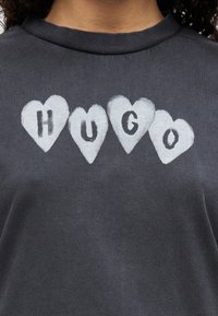 Γκρι ανθρακί μπλουζάκι με στρογγυλή λαιμόκοψη που φέρει τρεις λευκές καρδιές. Μέσα σε κάθε καρδιά είναι η λέξη "HUGO" με έντονα μαύρα γράμματα.