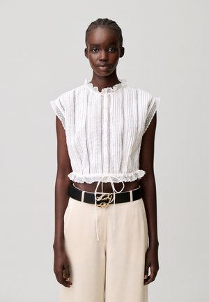 Mujer de piel oscura con el pelo trenzado que lleva una blusa blanca sin mangas con encaje y pantalones anchos color crema con cinturón negro.