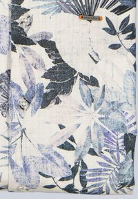 Light-colored fabric with blue and black tropical leaf patterns, showing buttons and a small rectangular metal brand tag.