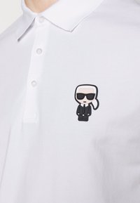 Bílá polo košile vyrobená z bavlny, na levé straně hrudi s nášivkou kreslené postavičky v obleku a slunečních brýlích, která je černá a hnědá.