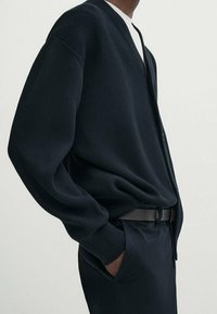 Marineblauwe geribbelde cardigan met een V-hals en lange mouwen. Draag het over een witte top, gecombineerd met donkere broeken en een zwarte riem.