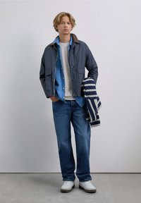 Outfit casual que presenta una chaqueta azul oscura con un collar marrón, superpuesta sobre una camisa azul clara, combinada con jeans azules y zapatillas blancas.