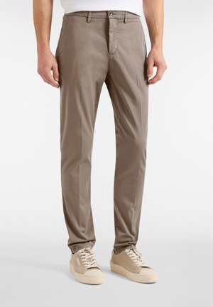Uomo con pantaloni dritti color taupe e sneakers beige, in piedi su uno sfondo bianco semplice.
