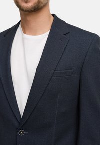 Mornarsko plavi sako uzorka riblje kosti s izrezanim reverom i jednom kopčom, u kombinaciji s bijelom majicom s okruglim izrezom ispod.